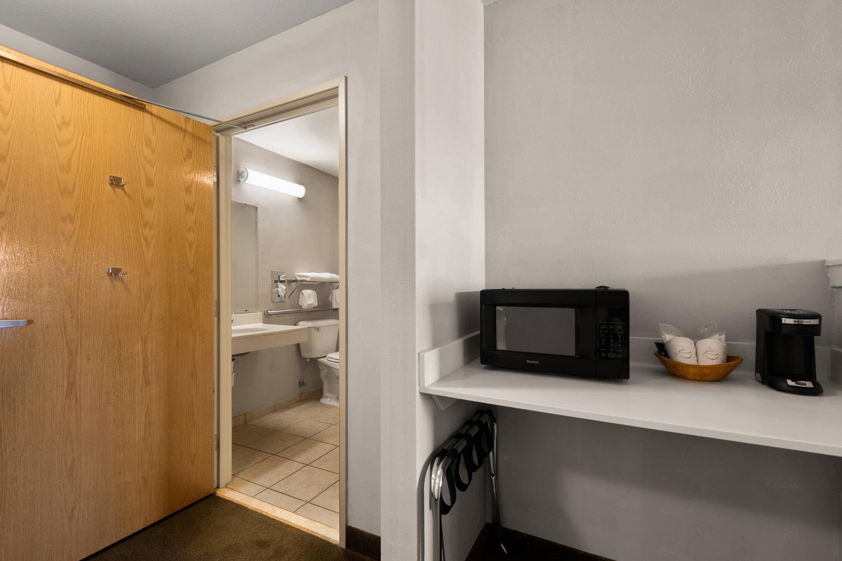 A hotel room with a wooden door, a microwave on a shelf, a coffee maker, and a small bowl with items.