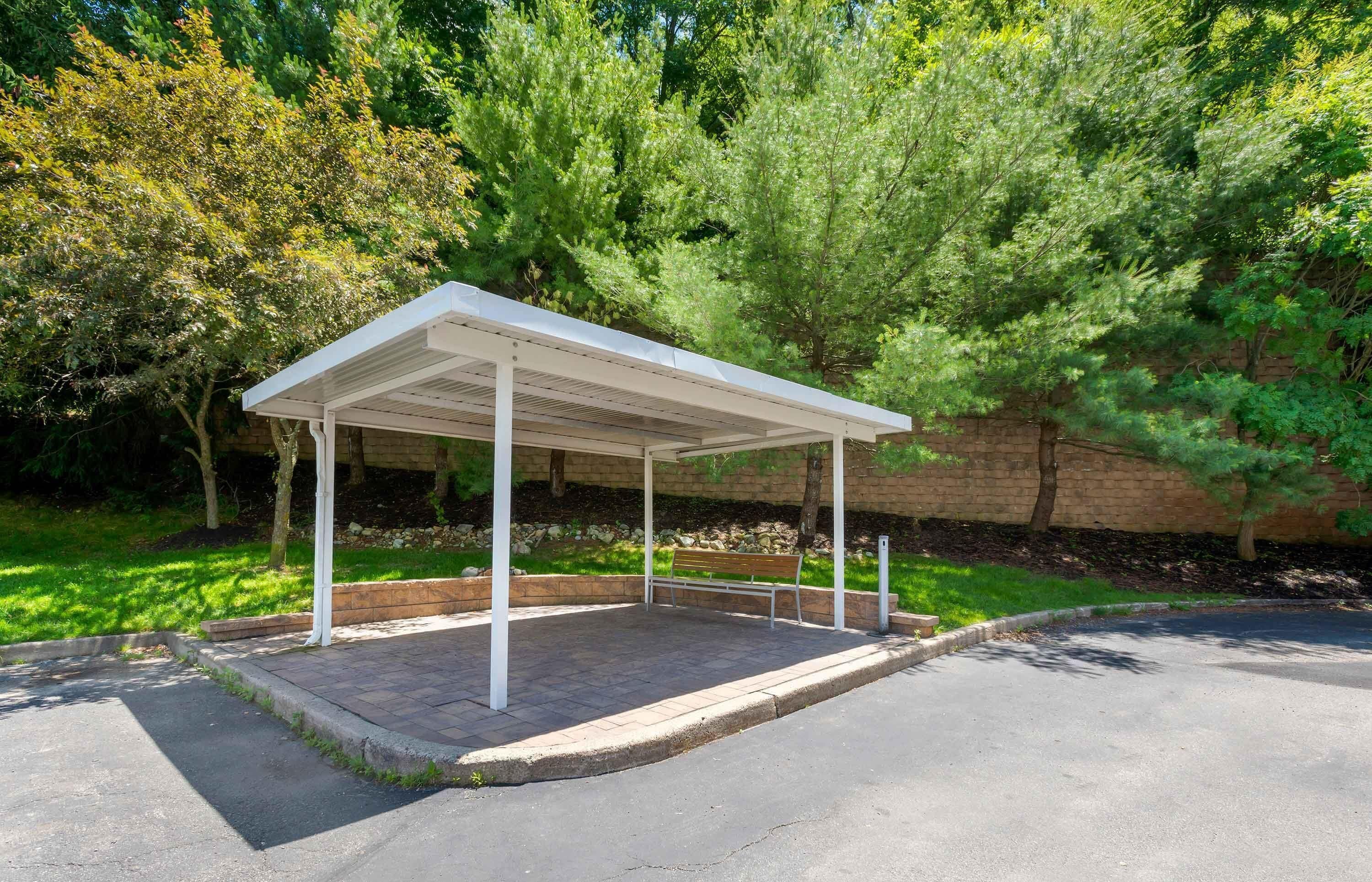 A covered outdoor seating area with a bench, surrounded by trees and a brick wall.