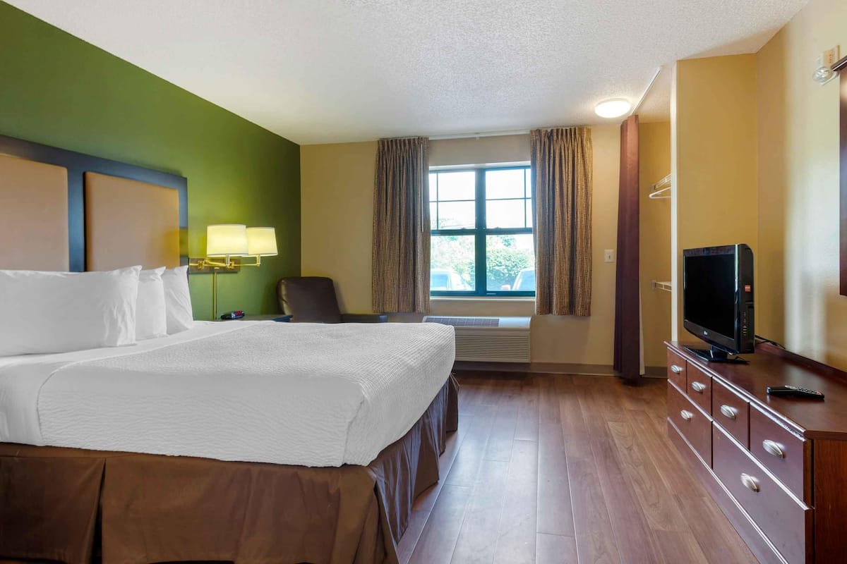 Une chambre d’hôtel avec un grand lit, un bureau avec une télévision, une chaise et une fenêtre avec des rideaux.