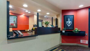 Un espace d’accueil d’hôtel avec un mur rouge, un comptoir d’accueil, un téléphone, un écran numérique et un panneau indiquant la sortie.