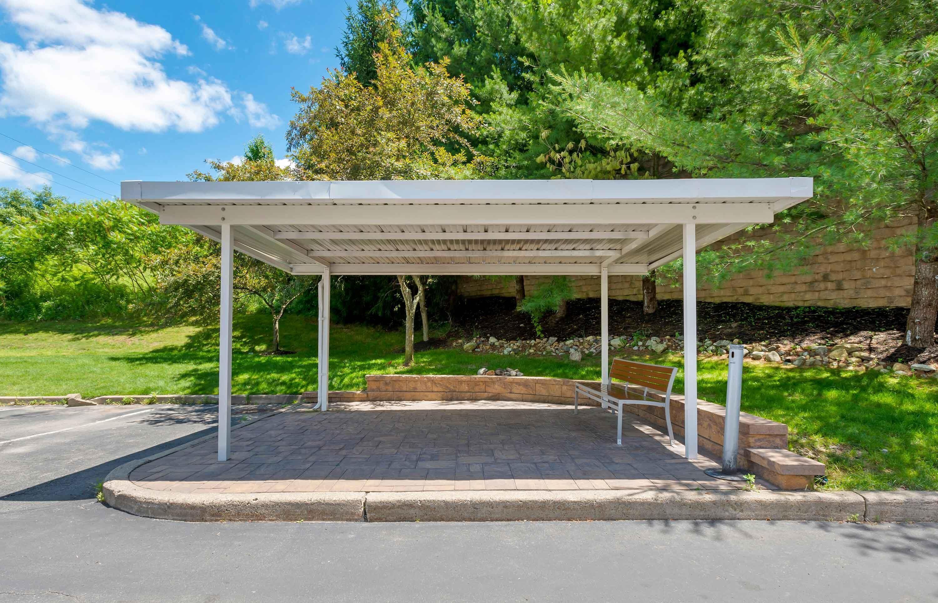 A covered outdoor seating area with a bench, surrounded by greenery and a stone wall.