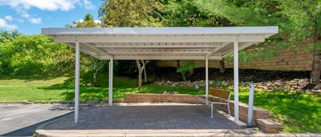 A covered outdoor seating area with a bench, surrounded by greenery and a stone wall.