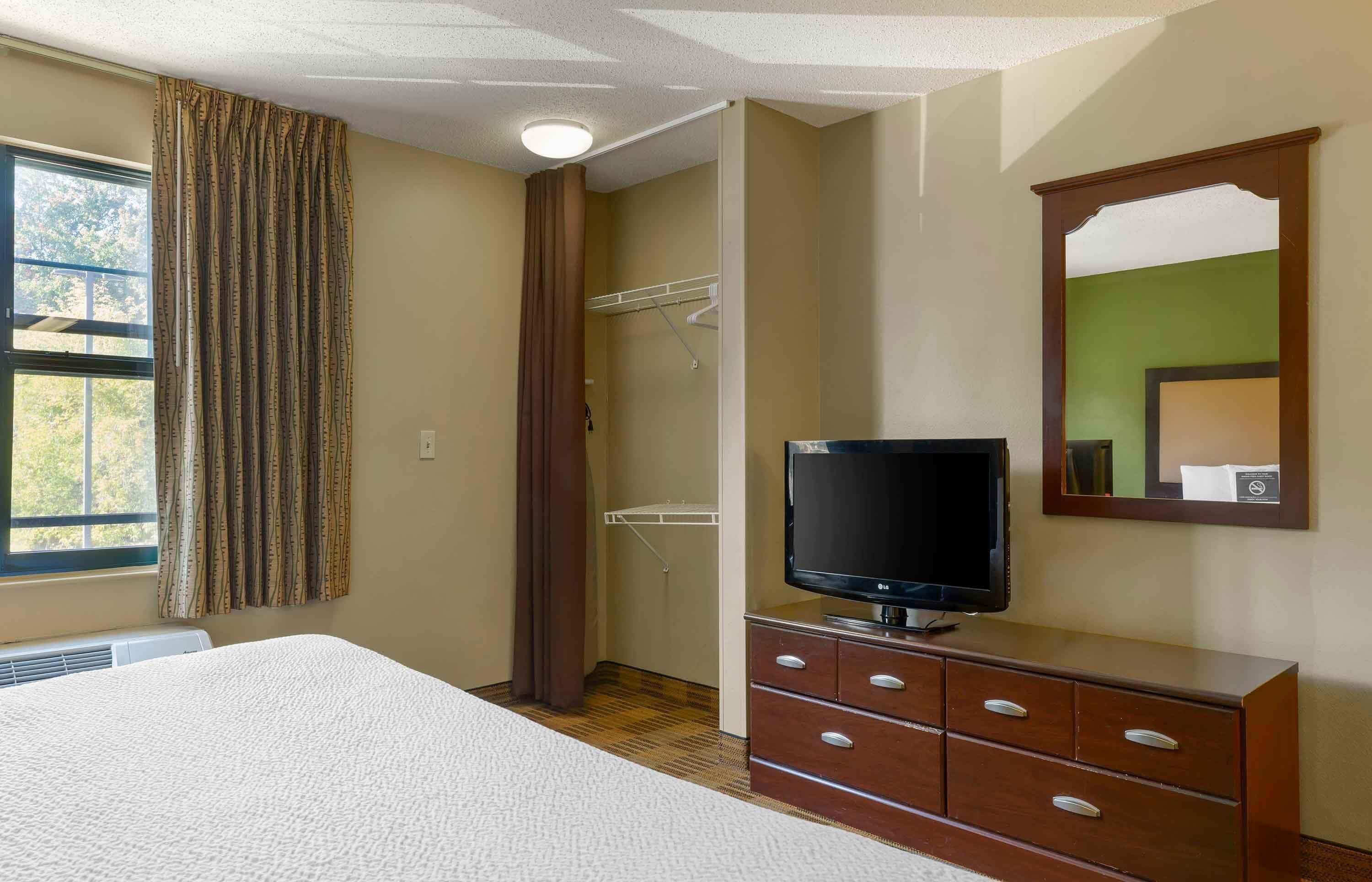 A hotel room with a bed, a TV on a wooden dresser, a closet with a hanging rack, and a window with curtains.