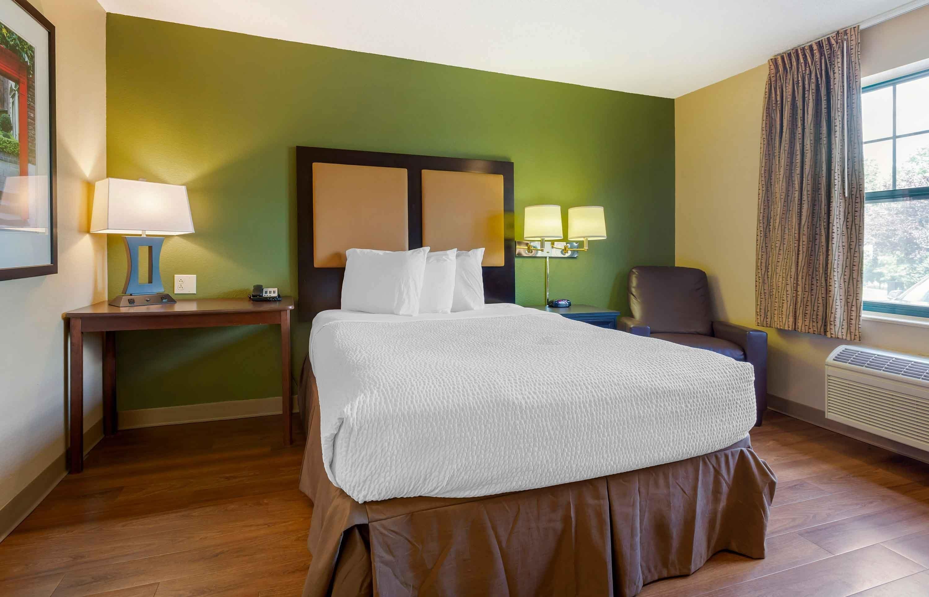 Ein Hotelzimmer mit einem großen Bett, einem Nachttisch mit Lampe, einem Sessel und einem Fenster mit Vorhängen.