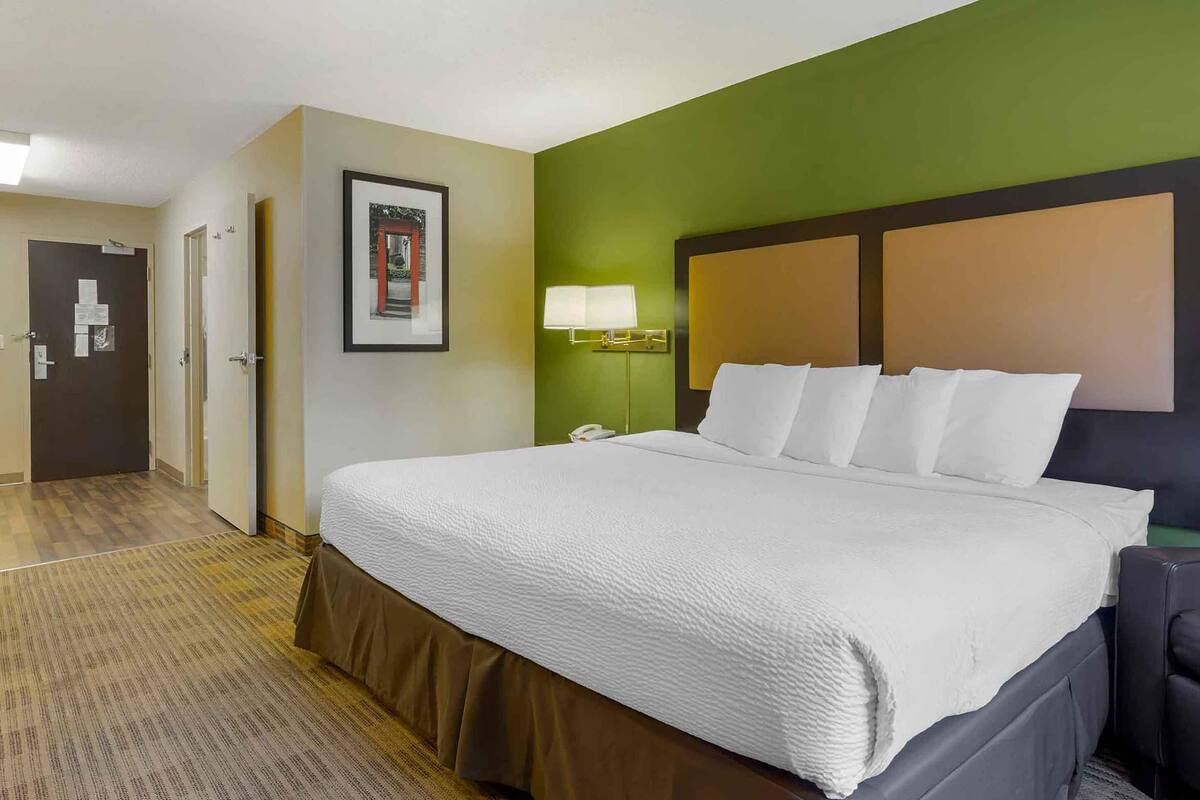 Une chambre d’hôtel avec un grand lit, une table de chevet avec une lampe, un tableau encadré au mur et une porte sur laquelle est affiché un avis.
