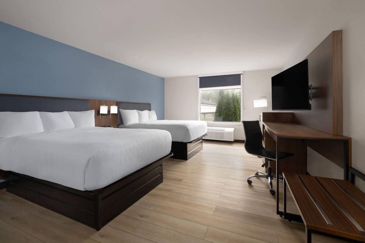 A hotel room with two beds, a desk, a chair, a TV, and a window with a view of trees.