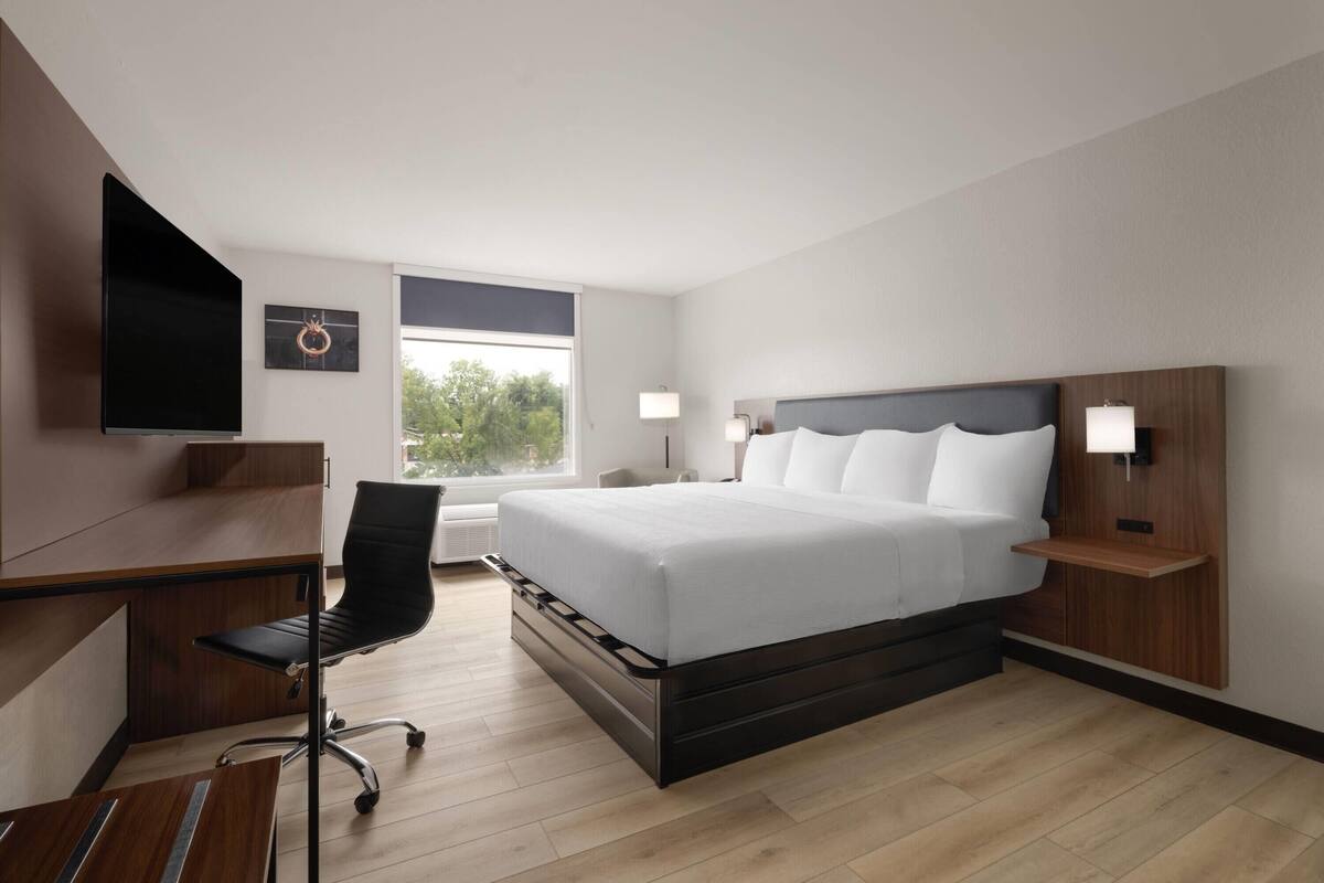 A modern hotel room with a large bed, a desk, a chair, a flat-screen TV, and a window with a view of greenery.