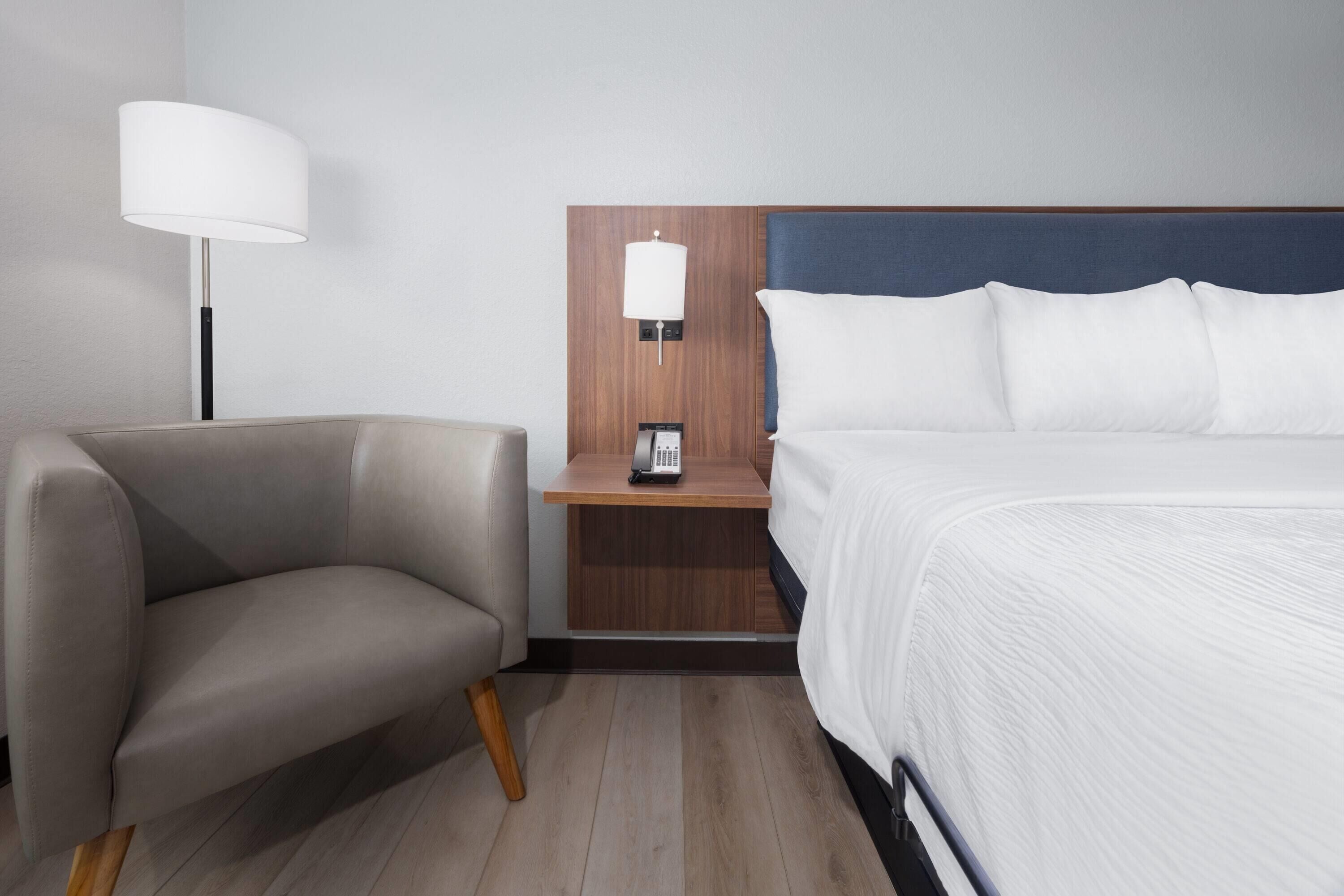A hotel room with a bed, a chair, a nightstand with a lamp, and a phone.