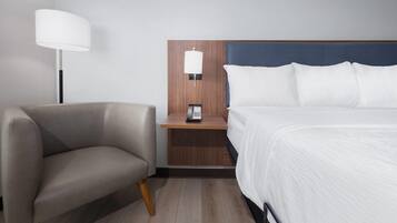 A hotel room with a bed, a chair, a nightstand with a lamp, and a phone.