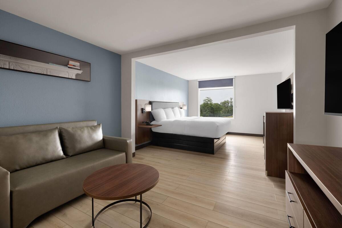 A modern hotel room with a sofa, a round coffee table, a bed with a headboard, a TV, and a large window offering a view of the outdoors.