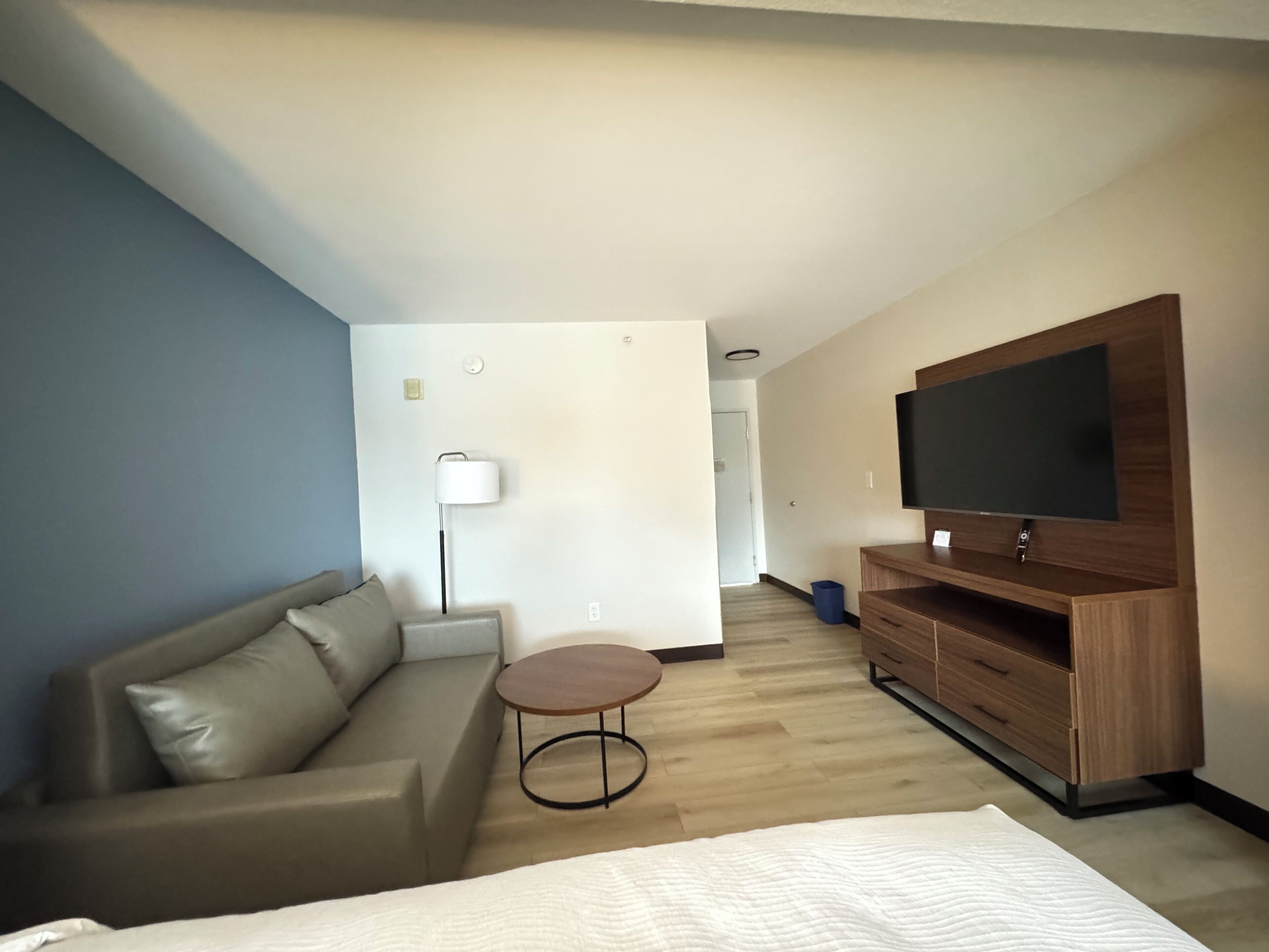 A modern hotel room with a bed, a sofa, a small round table, a TV on a wooden stand, and a lamp.