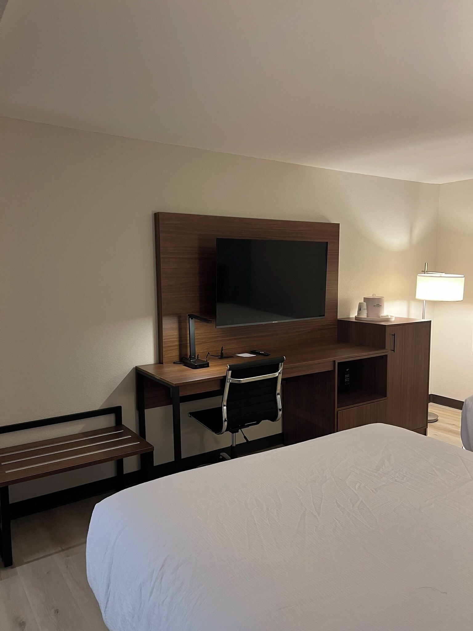 A hotel room with a large bed, a desk with a flat-screen TV, a chair, a lamp, and a bench.