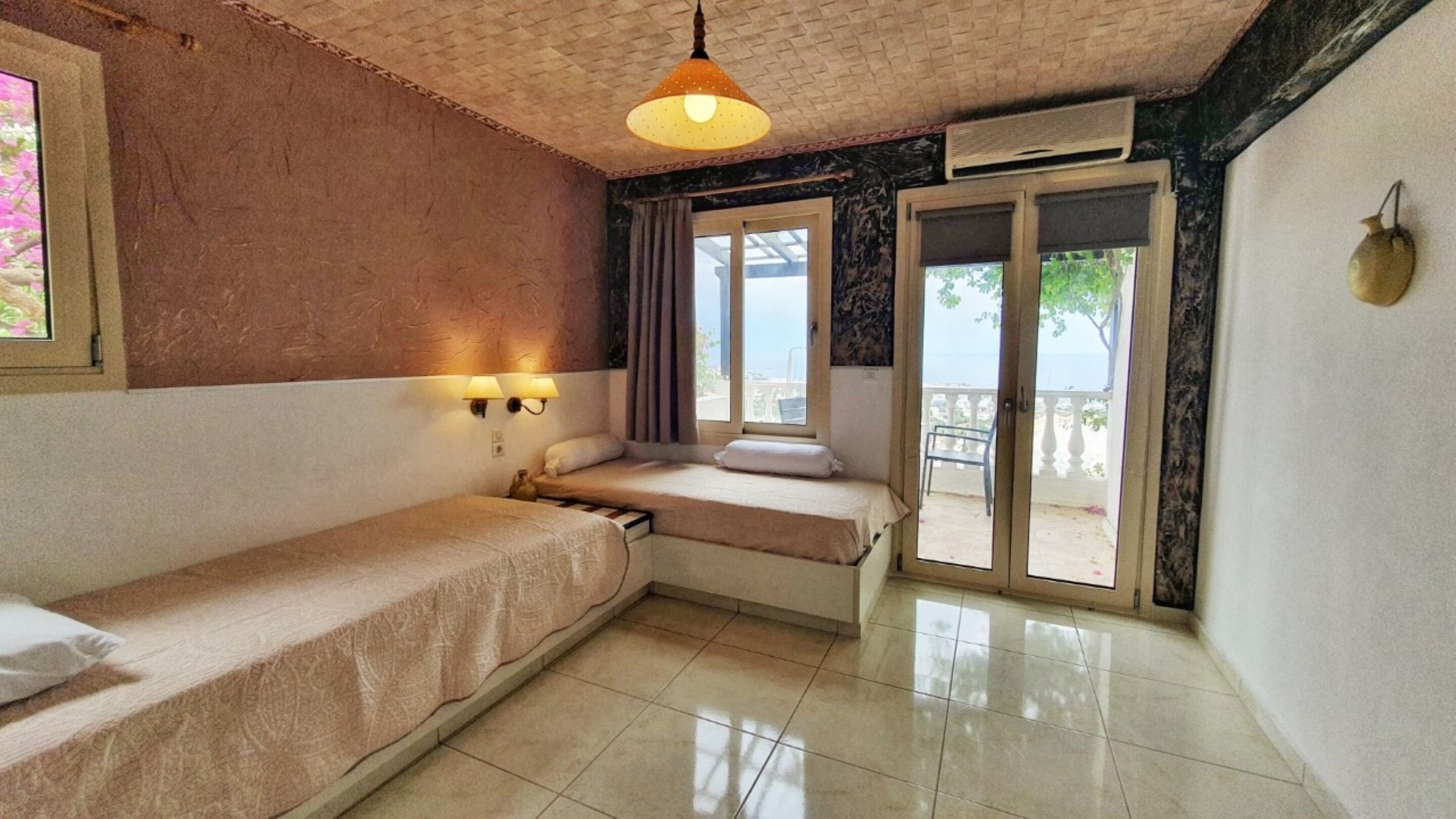 A small, compact room with two single beds, a window with curtains, a balcony, and a hanging light fixture.