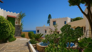 Un bâtiment situé sur la côte, avec un chemin de pierre menant à une piscine, le tout entouré d’une végétation luxuriante et sous un ciel bleu clair.