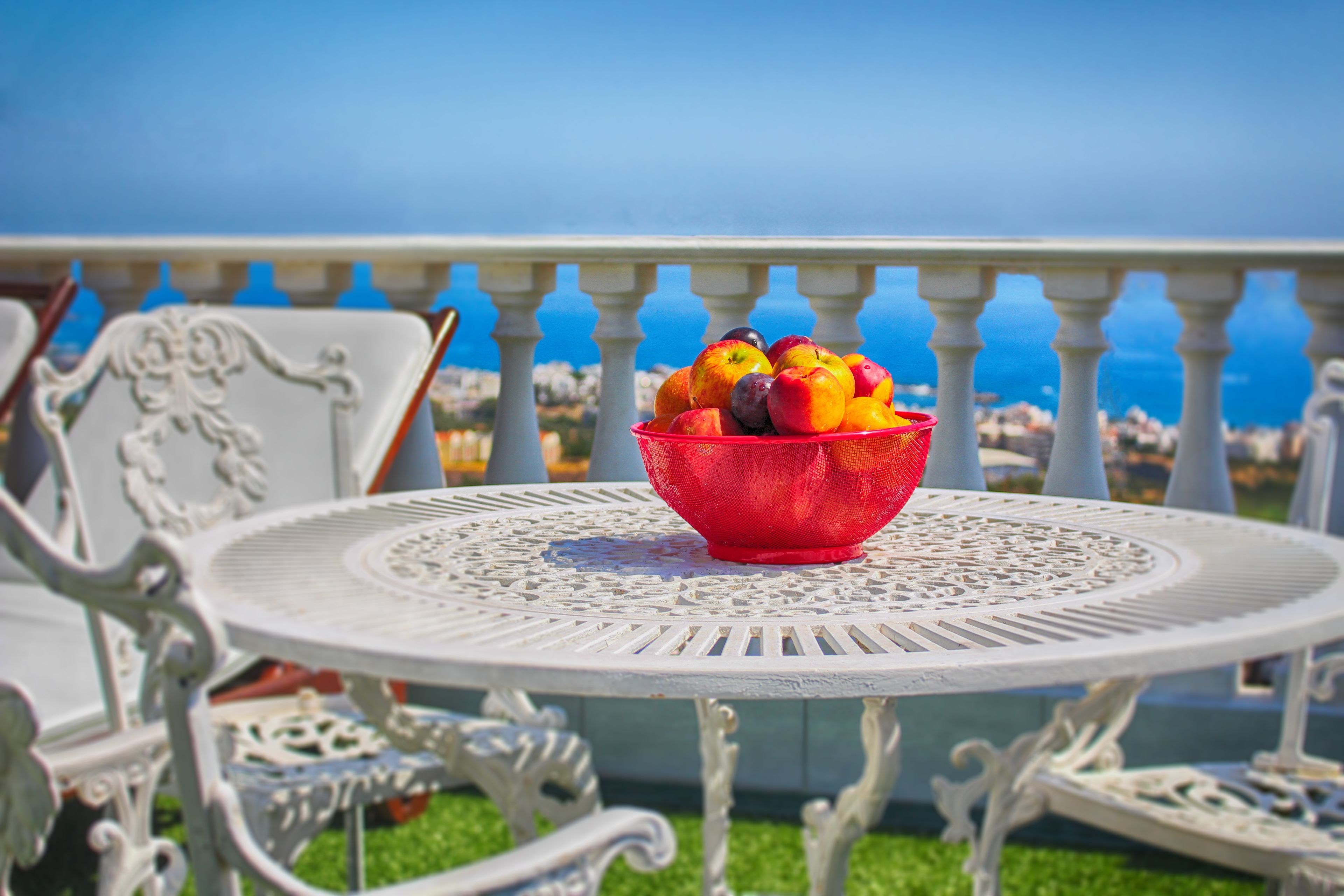 A round table with a red bowl of fruit, white ornate chairs, and a balcony with a view of the sea.