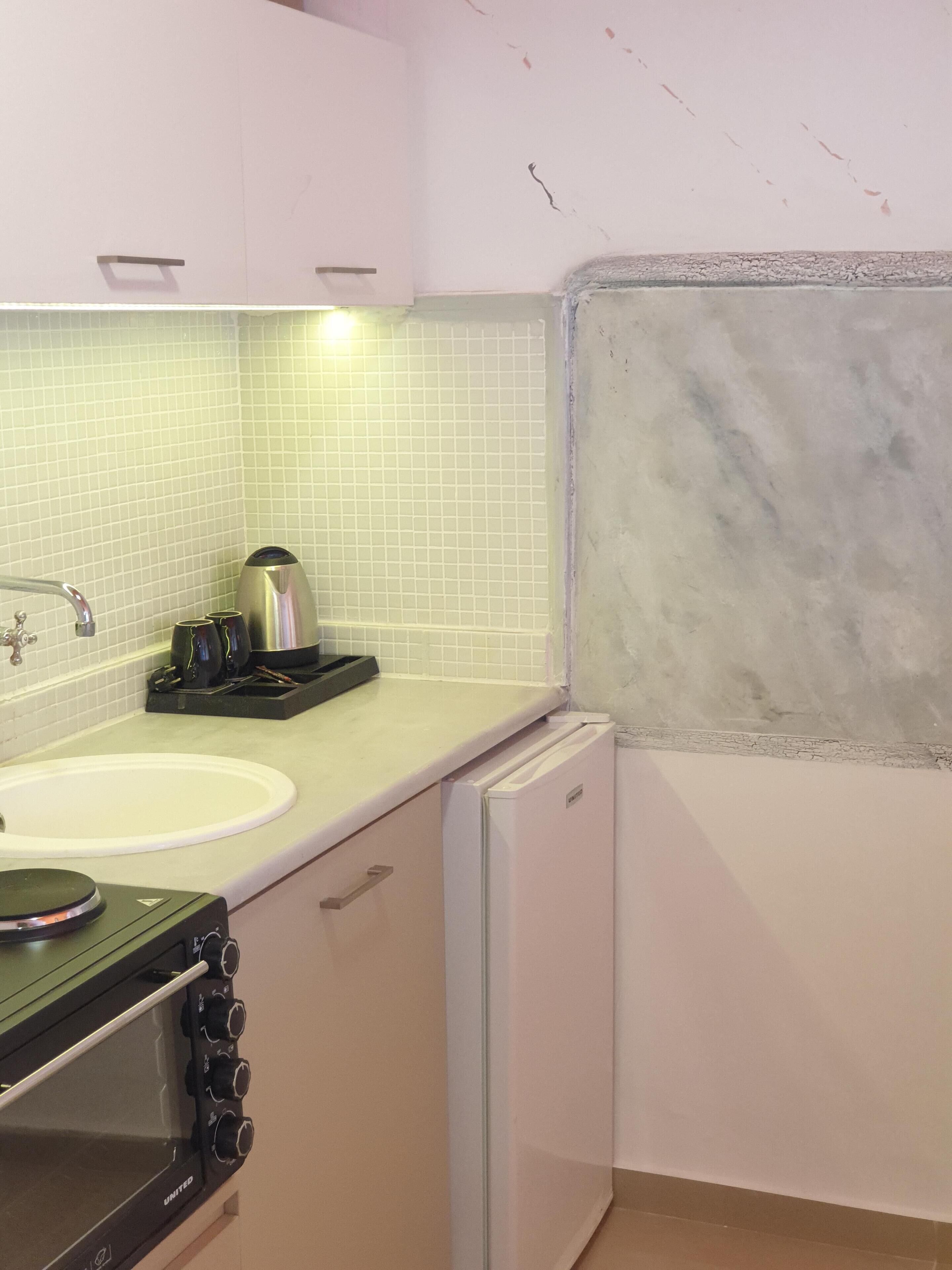 Een keuken met een witte spoelbak, een klein fornuis, een waterkoker en een compacte koelkast.