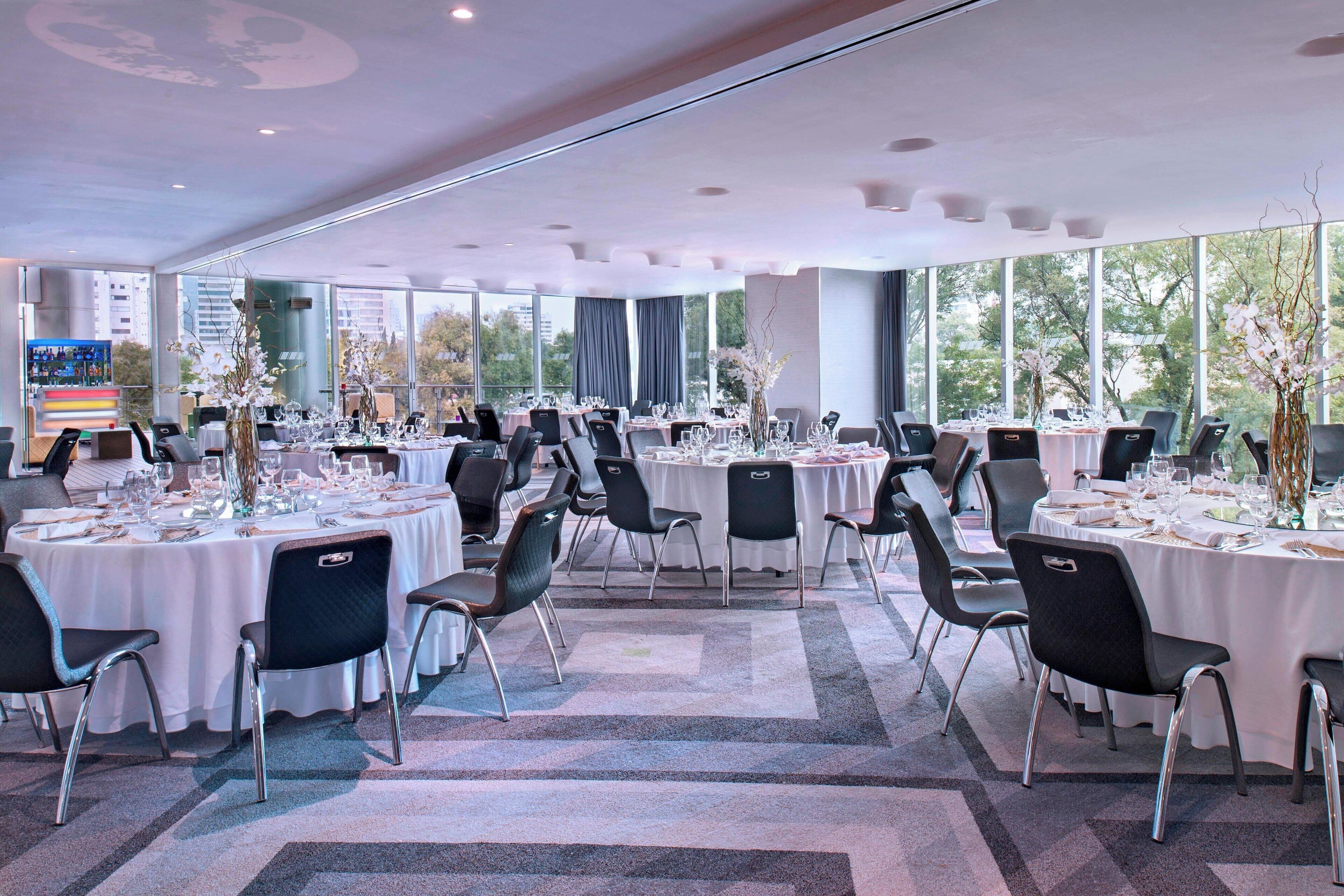 Una amplia sala de conferencias con varias mesas dispuestas para comer, grandes ventanales con vista a la ciudad y una decoración moderna.