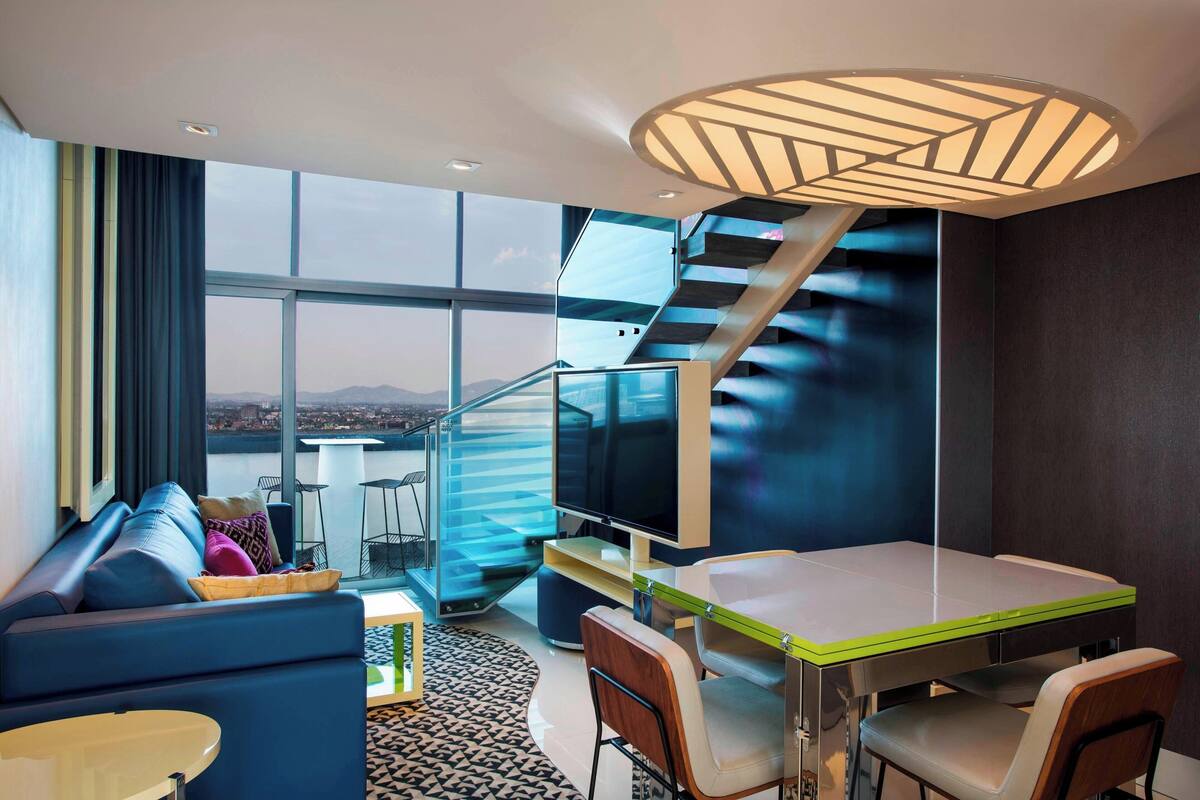 Una habitación de hotel moderna con un ventanal grande, una escalera, un área de comedor y un sofá.