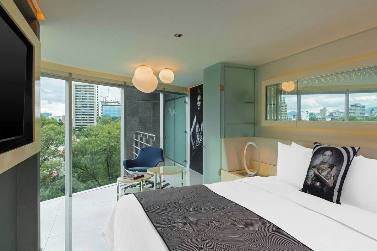 Una habitación de hotel moderna con una cama grande, vistas a la ciudad, una silla azul y una pequeña mesa.