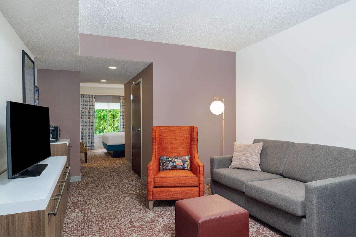 A hotel room with a television, a grey sofa, an orange armchair, a red ottoman, and a hallway leading to a bathroom.