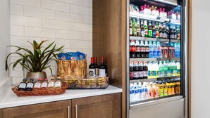 A kitchen counter with a refrigerator stocked with beverages, a plant, and packaged snacks. - Hilton Garden Inn Montgomery East (Montgomery)