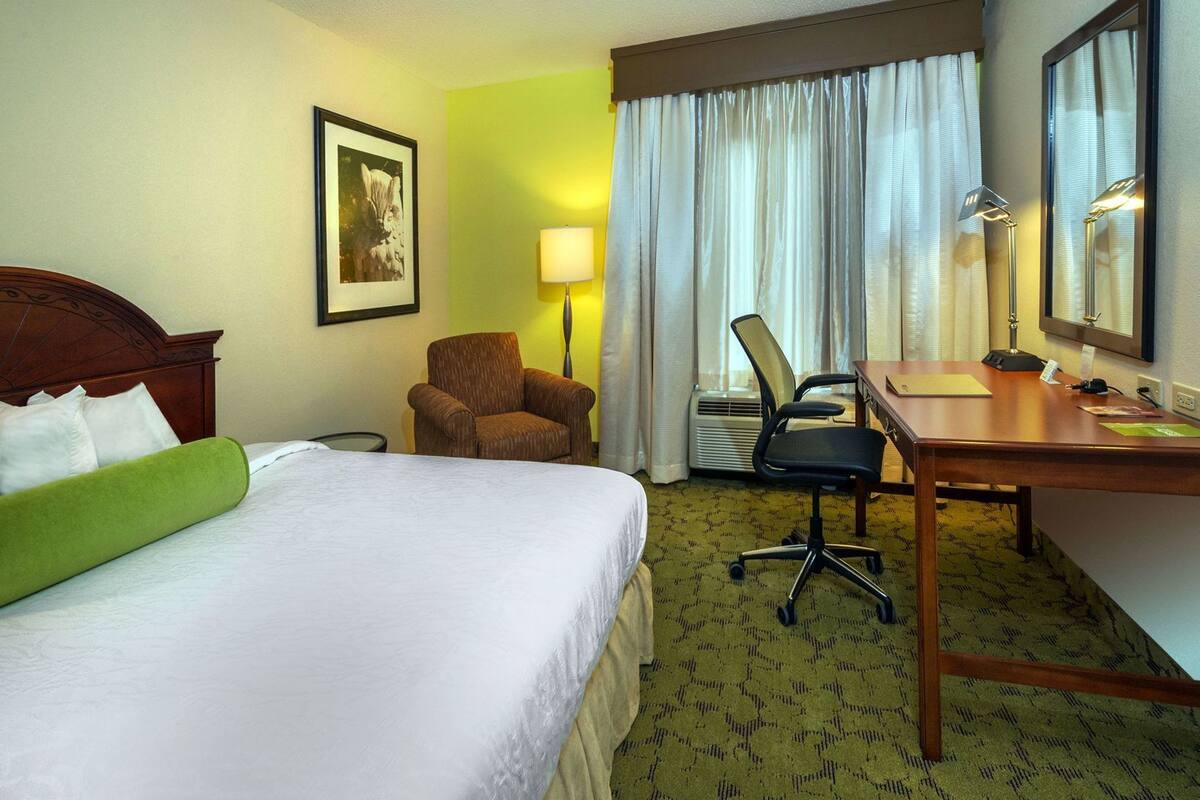 A hotel room with a bed, a desk, a chair, a lamp, and a picture on the wall.