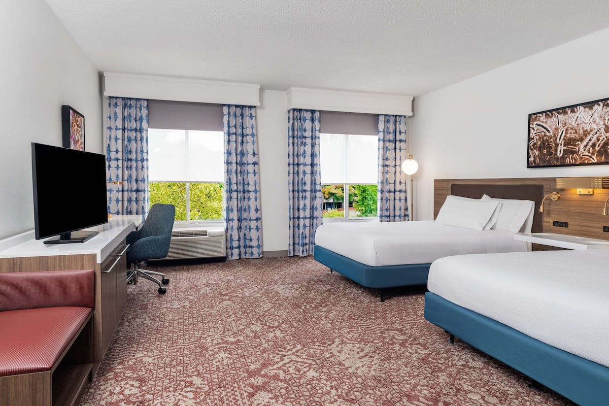 A hotel room with two beds, a desk with a TV, a chair, and a large window with curtains.