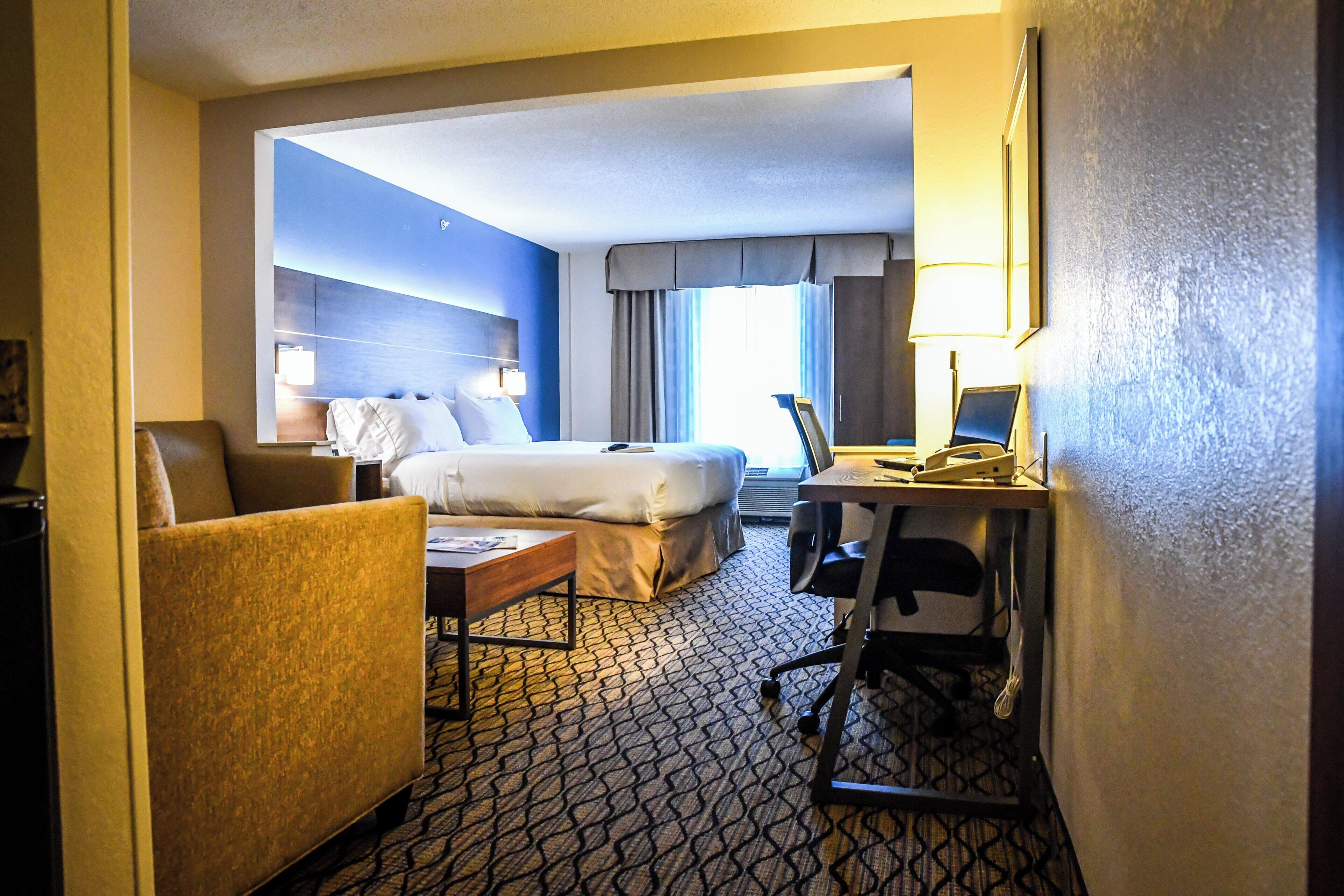A hotel room with a bed, a desk with a computer, a chair, and a small table.