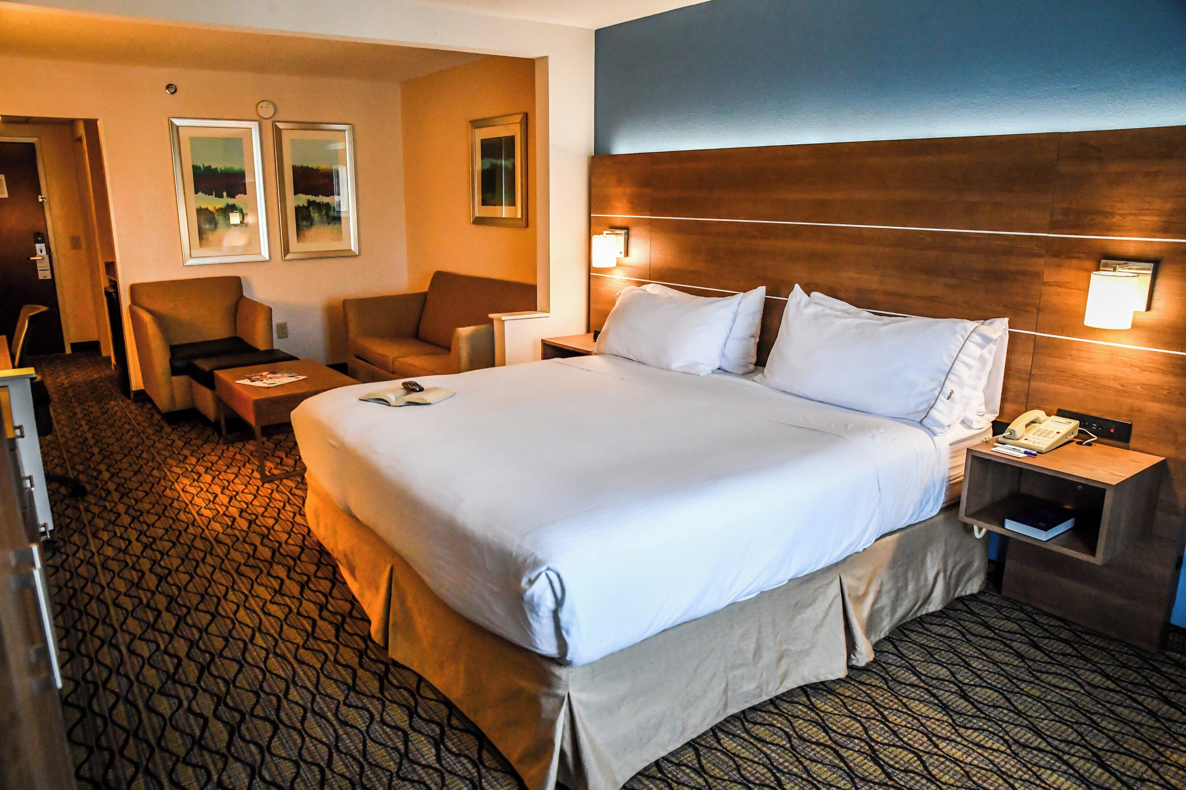 Habitación de hotel con una cama grande, dos sillones, una mesita y una mesita de noche con lámpara.