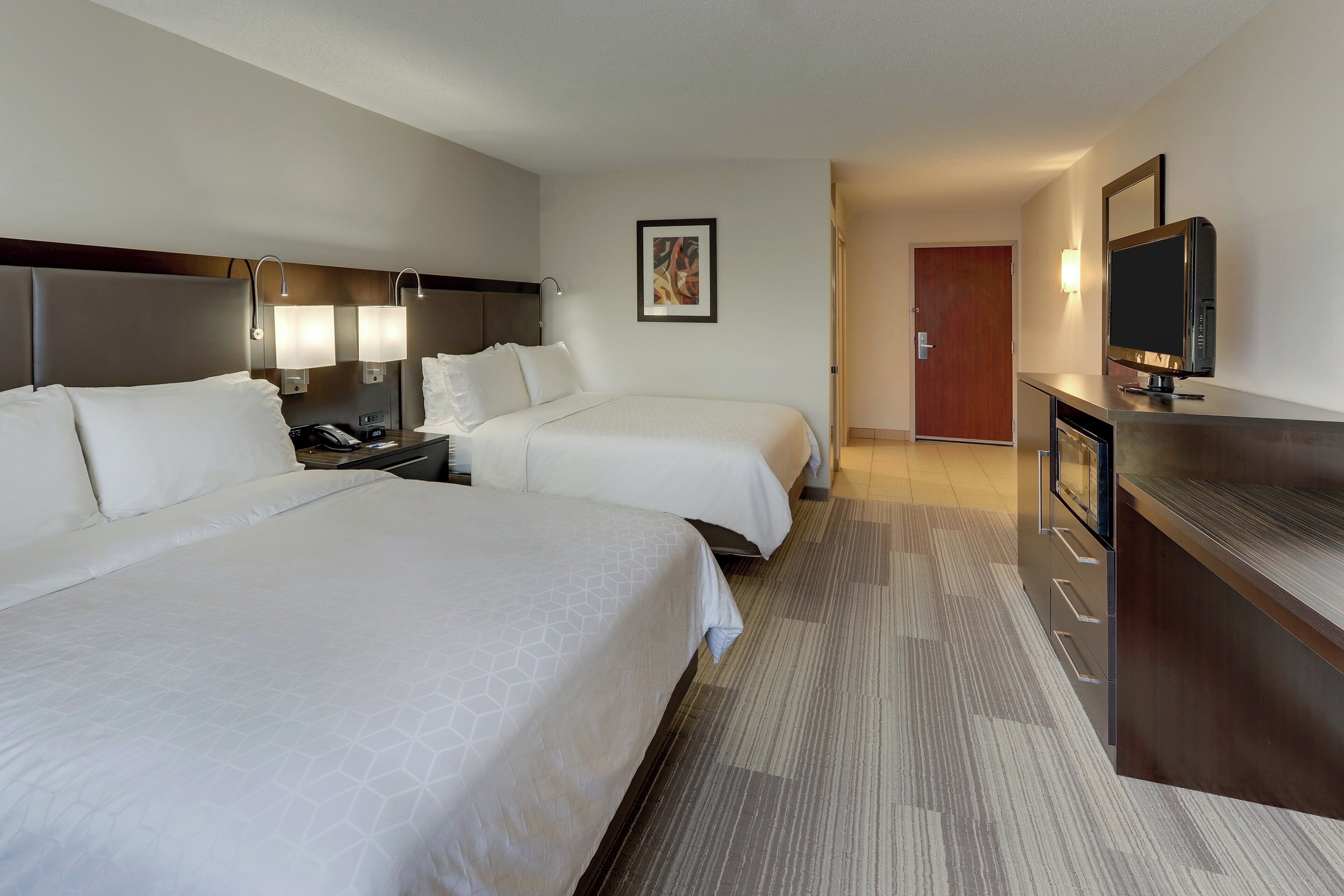 A hotel room with two beds, a desk with a television, a painting on the wall, and a door leading to another room.