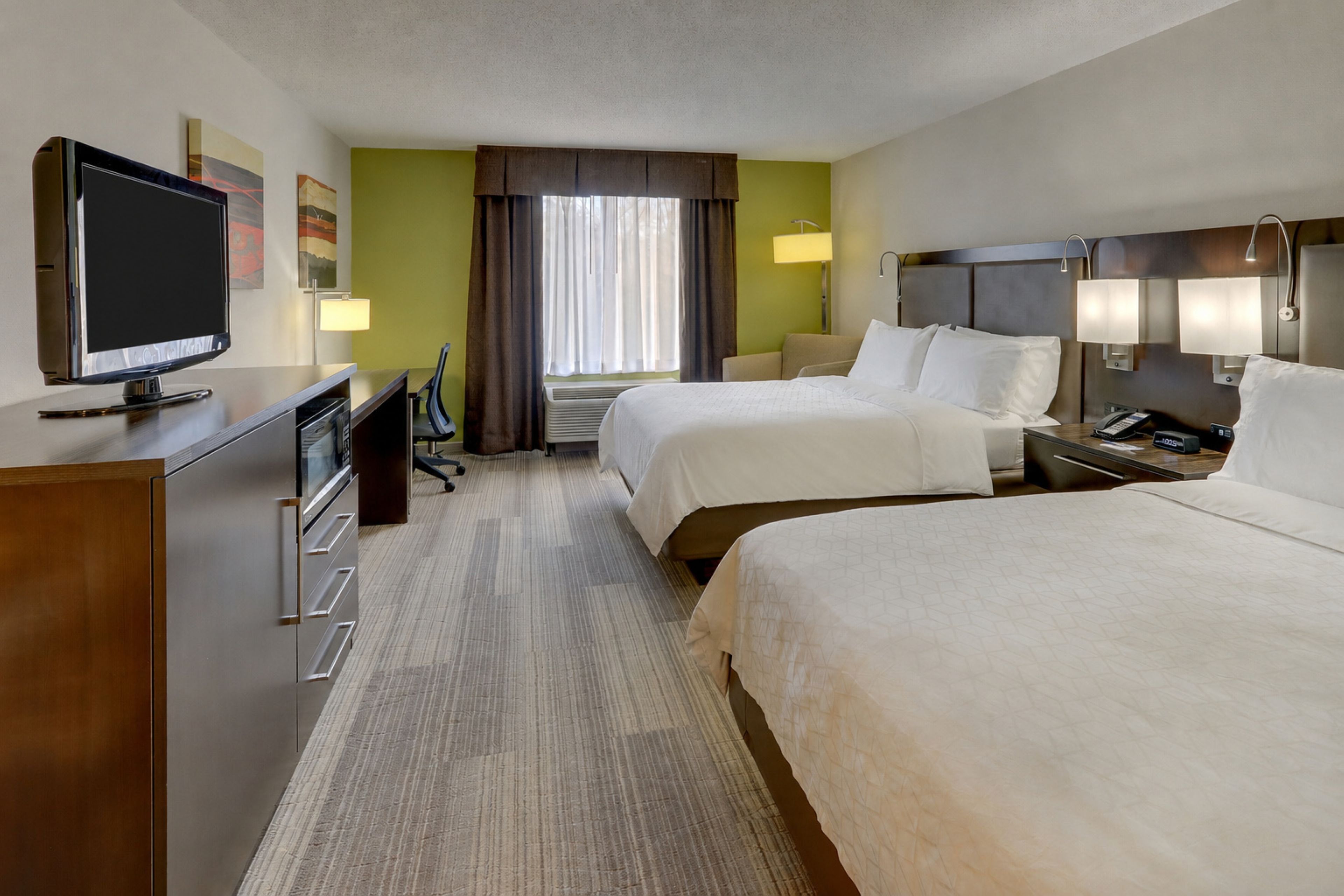 A hotel room with two beds, a desk with a television, a lamp, and a window with curtains.