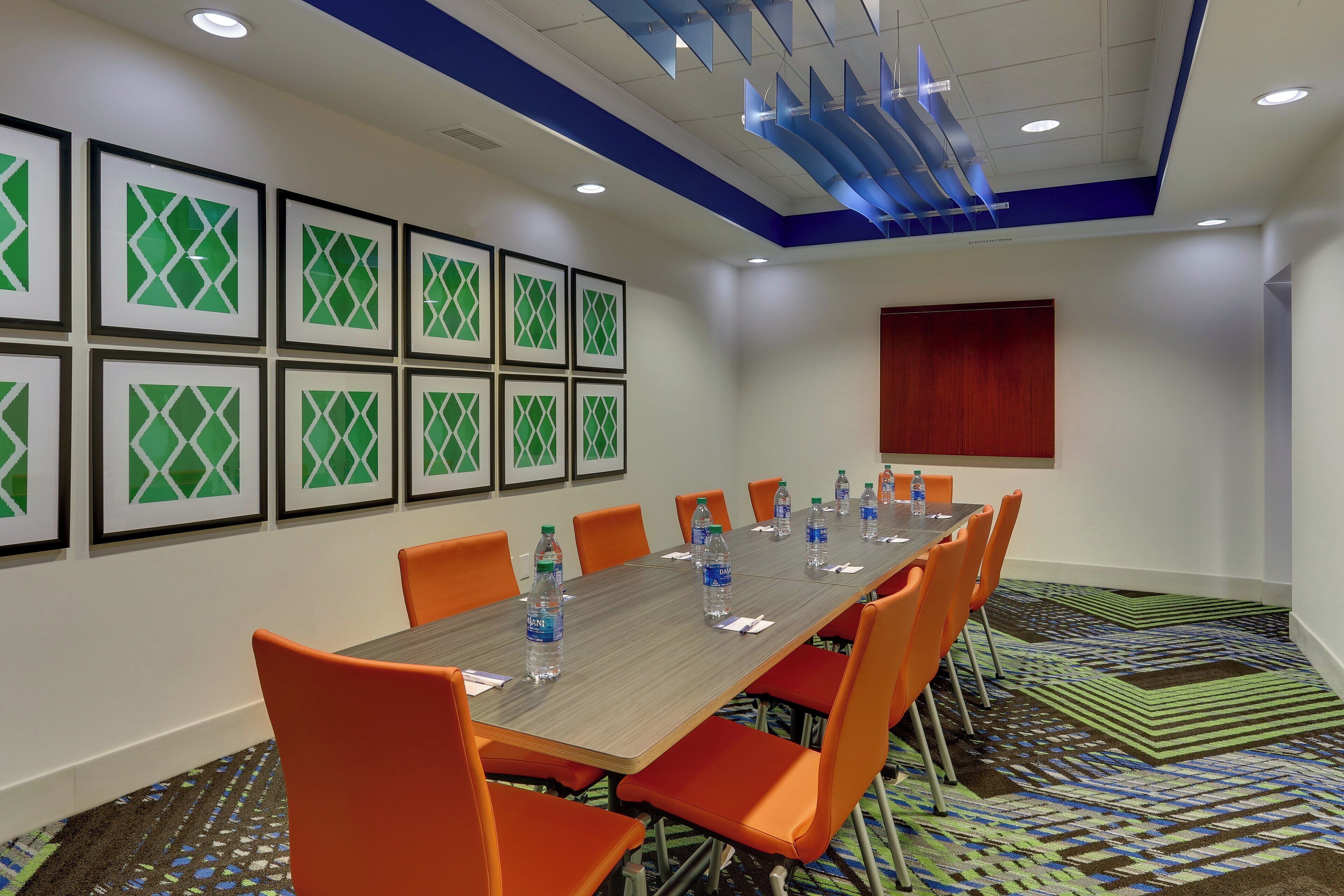 A conference room with a long table, orange chairs, framed pictures on the wall, and a patterned carpet.