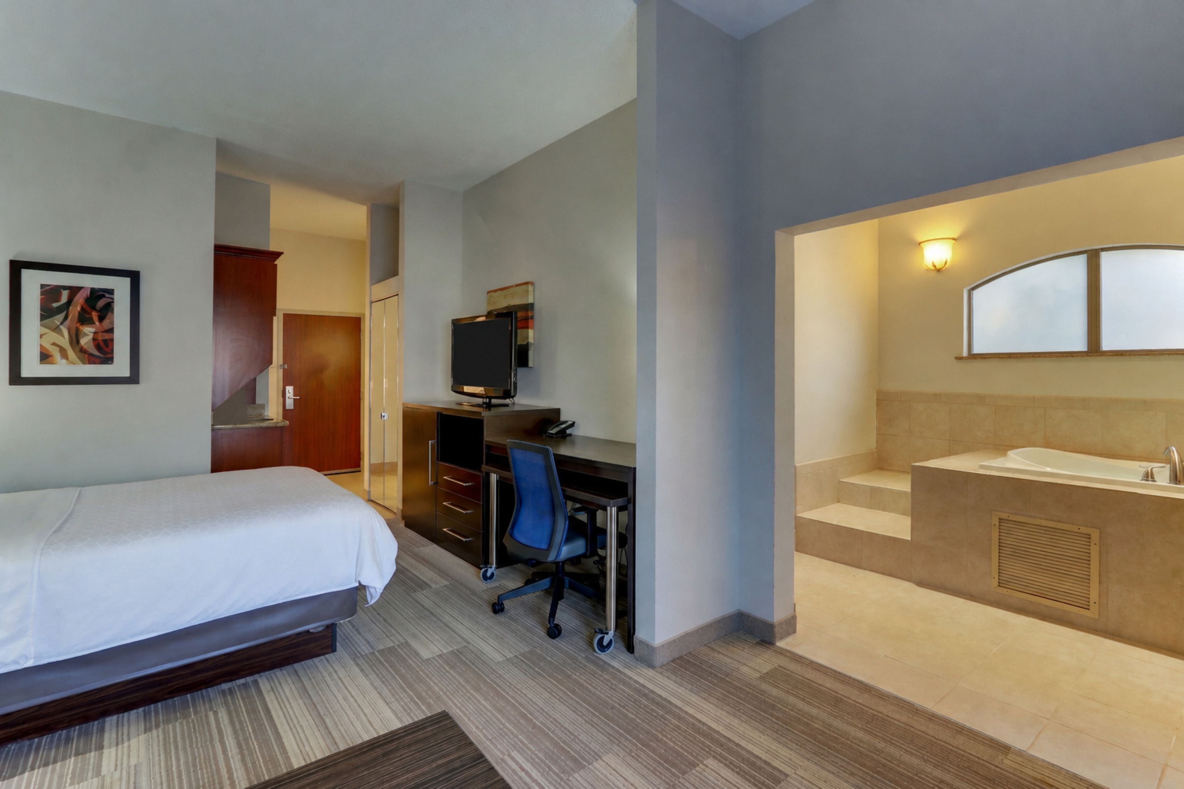 A hotel room with a bed, desk, chair, and a bathroom with a sink and mirror.