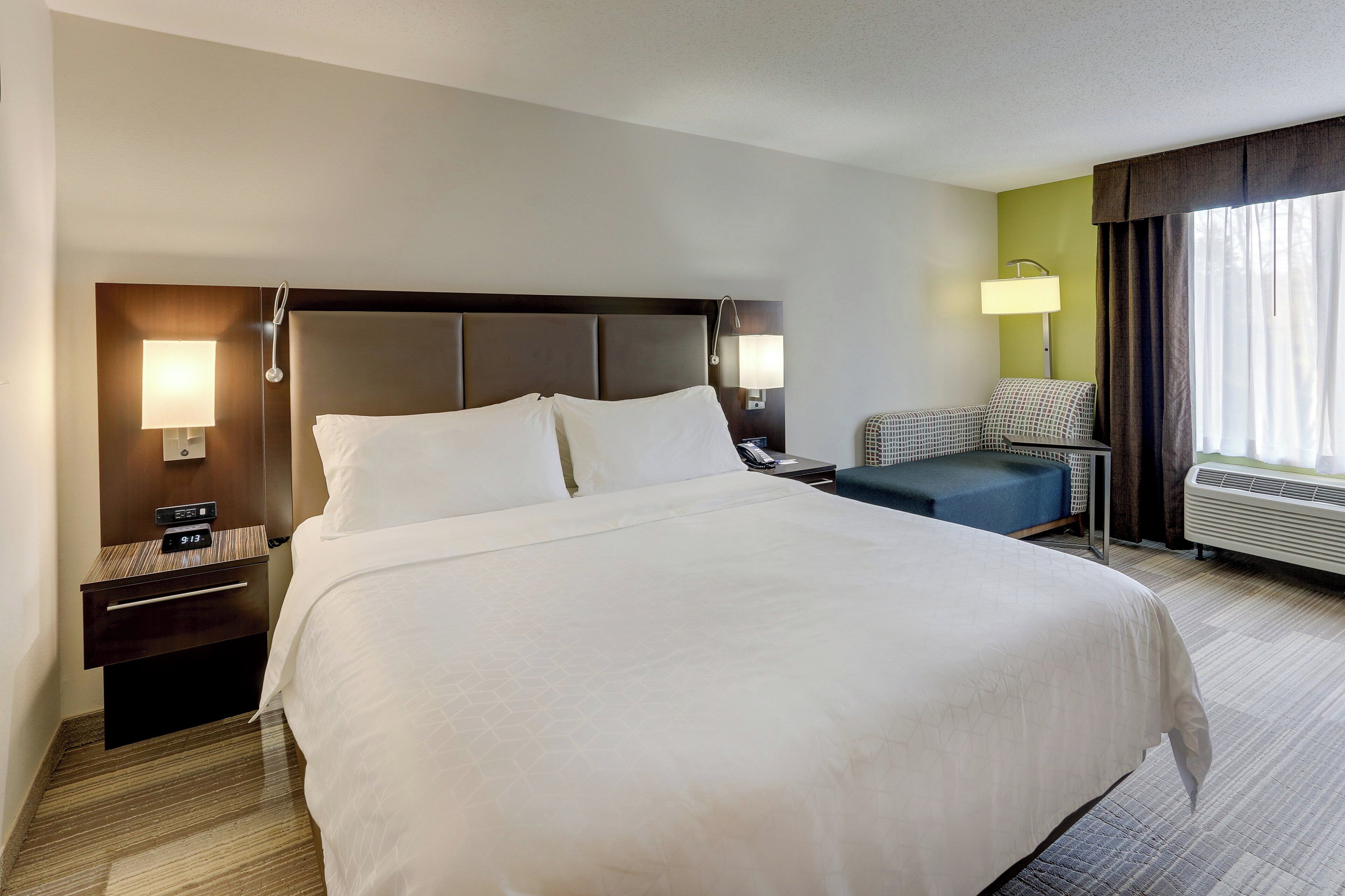 A hotel room with a large bed, bedside tables, a chair, a lamp, and a window with curtains.