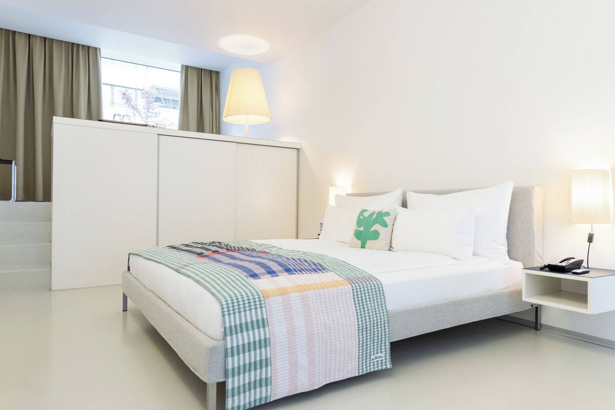 A modern hotel room with a neatly made bed, a bedside table with a lamp, and a large window with curtains.