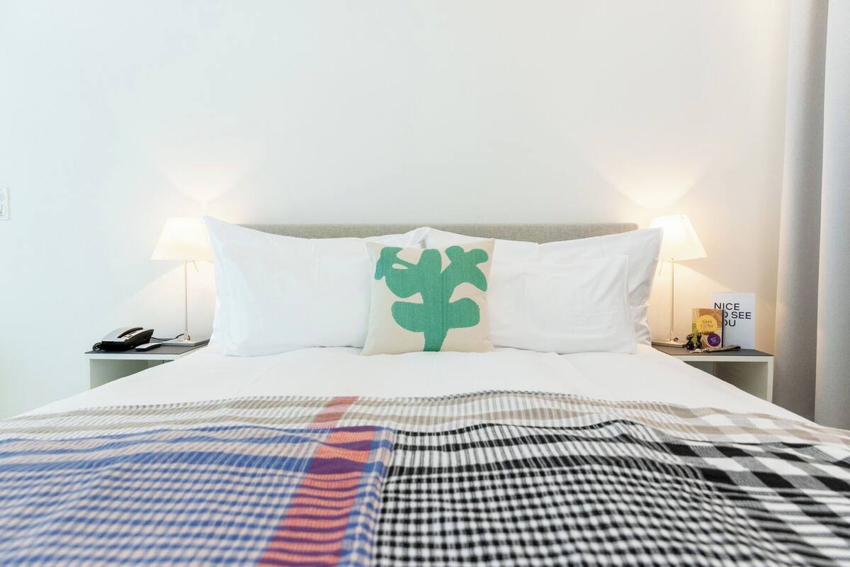 A neatly made bed with a checkered blanket, two bedside lamps, a nightstand with a phone, and a pillow with a green leaf design.