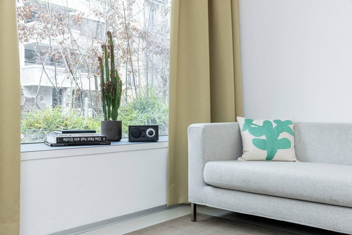 A modern living room with a grey sofa, a green cushion, a window with a view of trees and buildings, and a shelf with books and a radio.