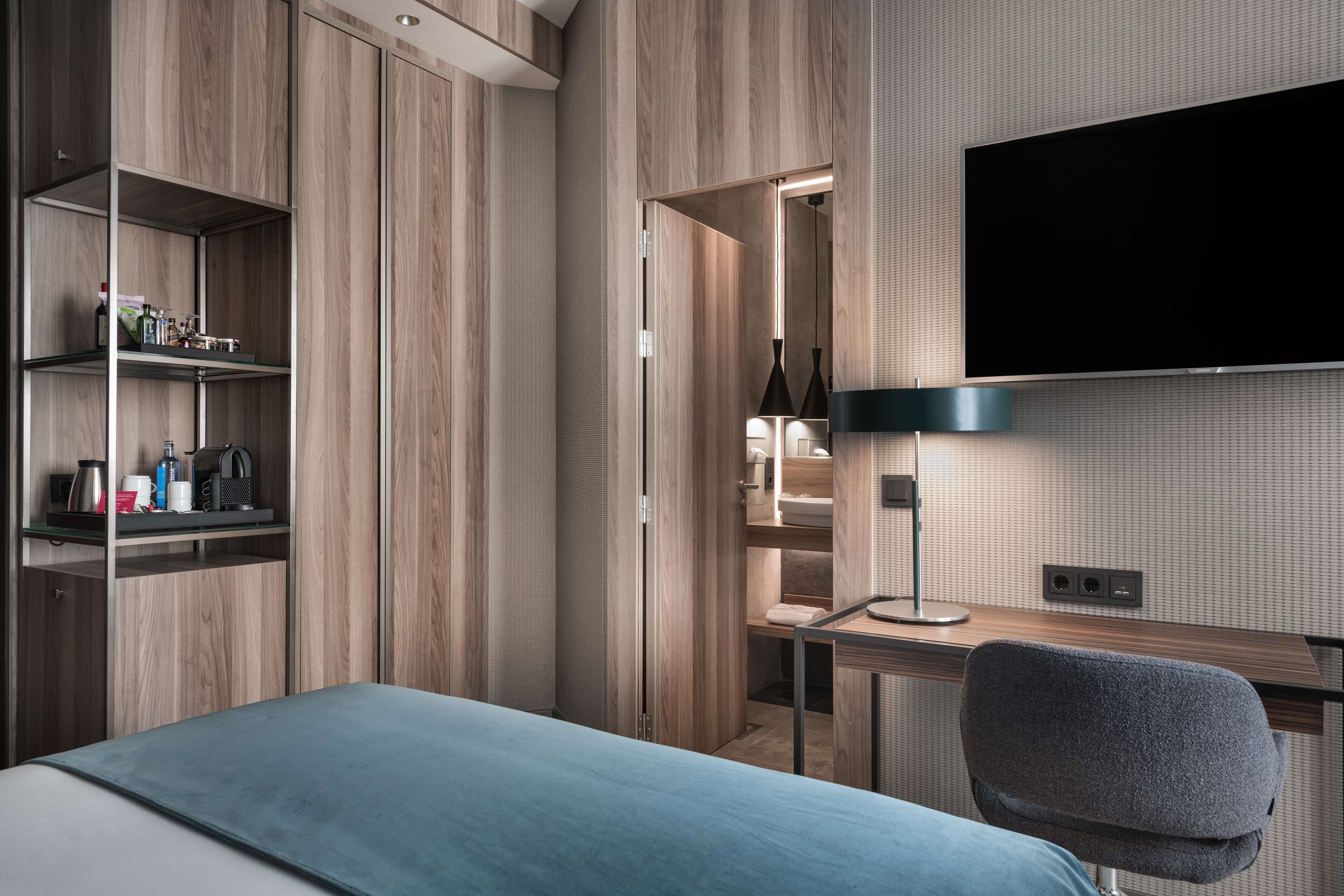 A modern hotel room with a bed, a desk with a computer, a TV, and built-in shelves with toiletries.