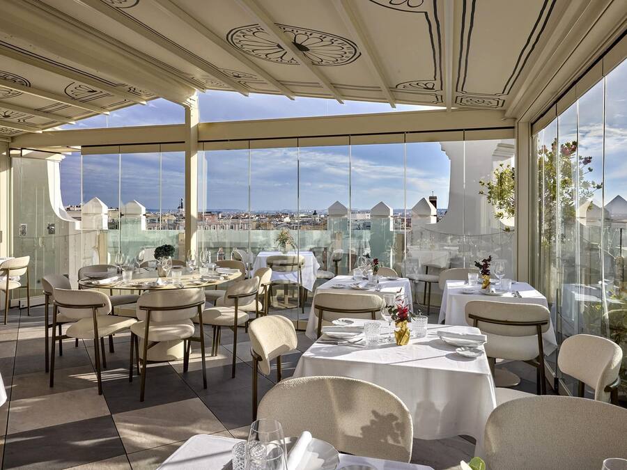 Um restaurante no terraço com mesas preparadas para refeições, grandes janelas com vista para a cidade e teto decorativo.