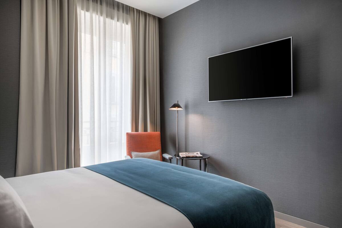 A modern hotel room with a large bed, a flat-screen TV mounted on the wall, a small side table, and a chair with a cushion.