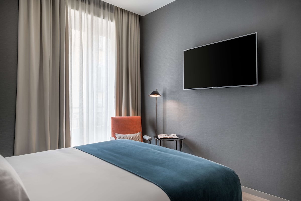 A modern hotel room with a large bed, a flat-screen TV mounted on the wall, a small side table, and a chair with a cushion.