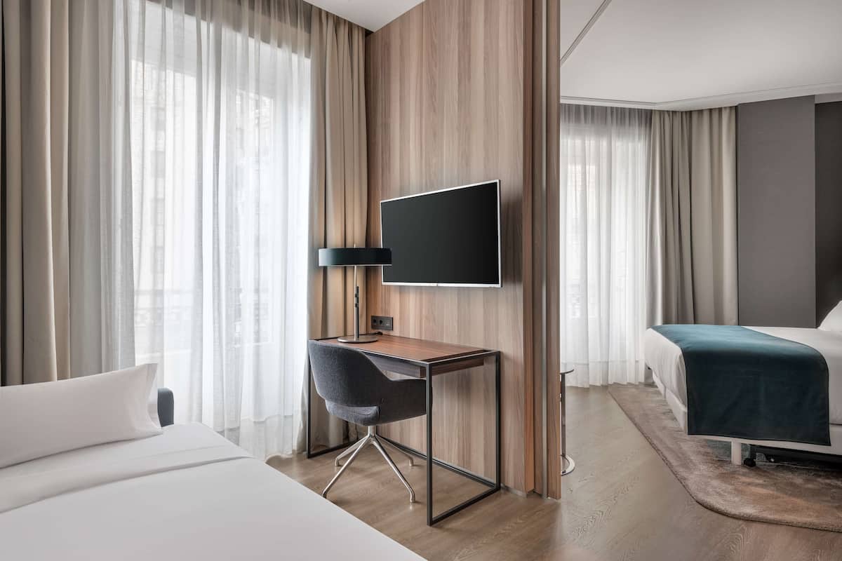 A modern hotel room with a bed, a desk with a chair, a television, and a large window with curtains.