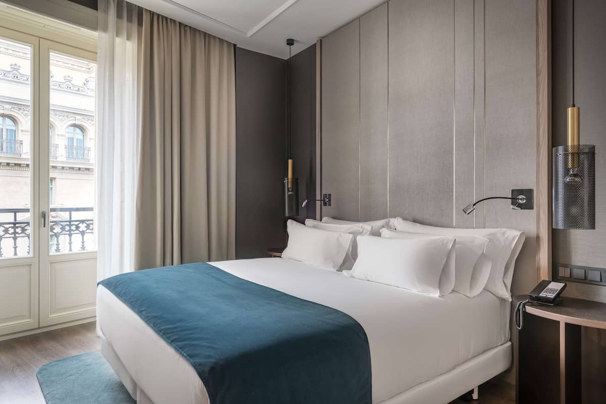 A hotel room with a large bed, a bedside table with a phone, and a view of a building through the window.