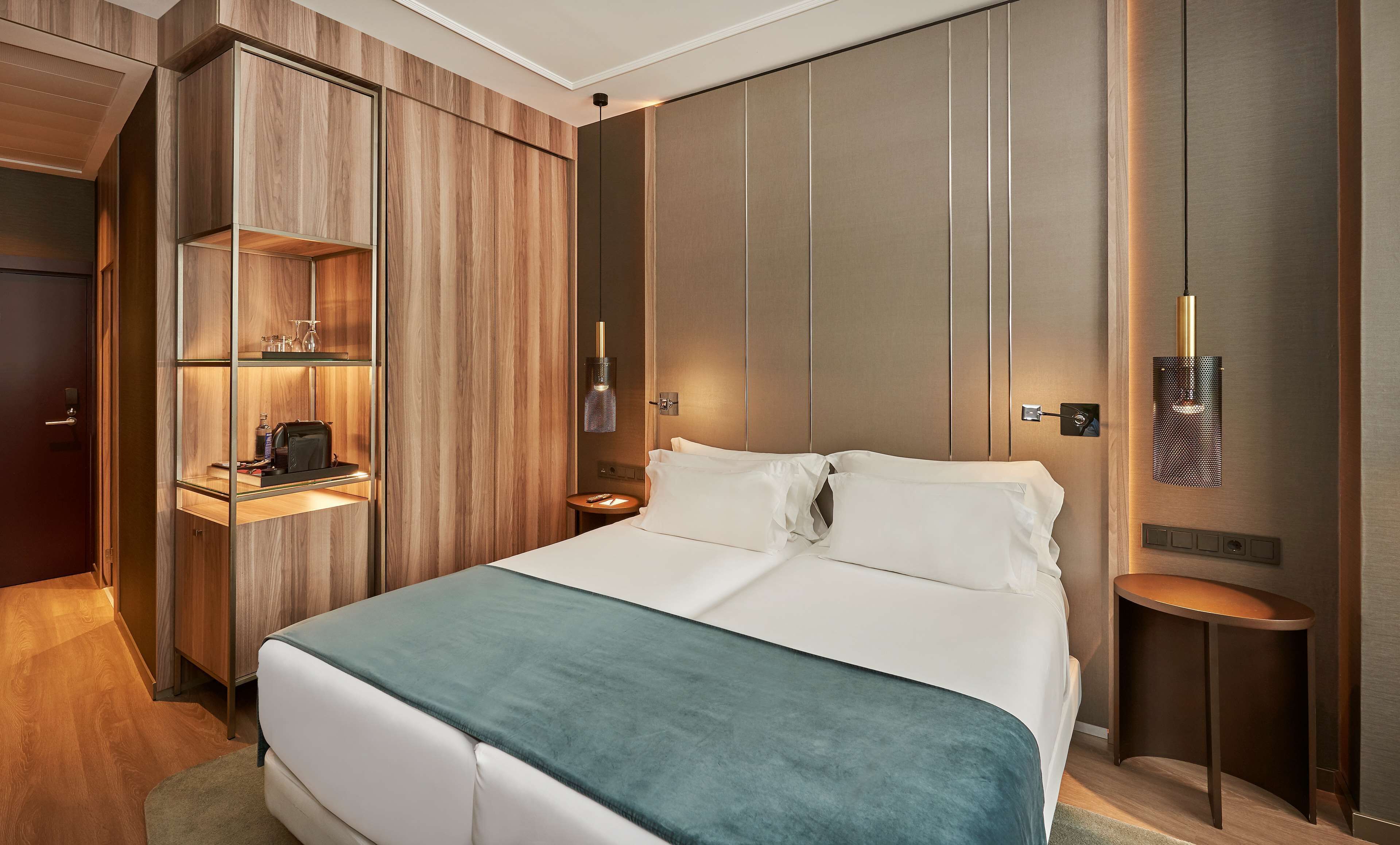 A modern hotel room with a large bed, wooden paneling, and built-in shelving with a minibar.