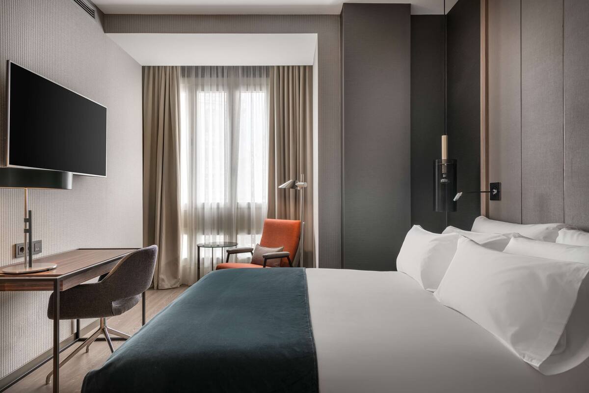 A modern hotel room with a large bed, a desk, a chair, a TV, and a window with curtains.