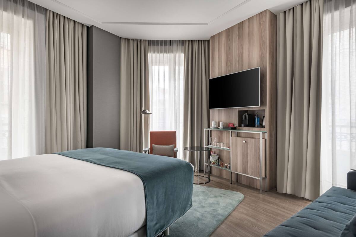 A modern hotel room with a large bed, a flat-screen TV, a desk with a coffee machine, and a seating area.