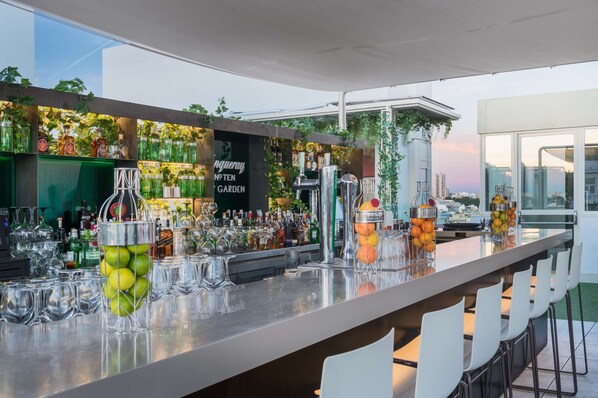 A bar area with a long counter, various bottles, and glasses. There are chairs along the counter. - NH Collection Madrid Gran Vía (Madrid)