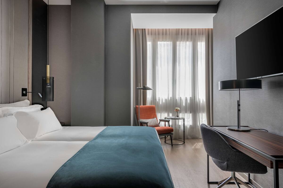 A hotel room with a bed, a desk, a chair, a lamp, and a view of the outdoors through a window with curtains.