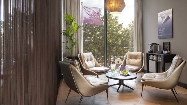 A modern lounge area with leather chairs, a round table, and a large window offering a view of trees and a flag. - Crowne Plaza London - Docklands by IHG (London)