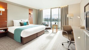 A hotel room with a large bed, a desk, a chair, a TV, and a view of a body of water. - Crowne Plaza London - Docklands by IHG (London)